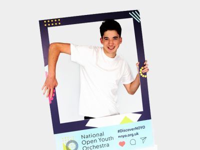 A boy wearing a white t-shirt stands in between a customised National Open Youth Orchestra Instagram frame, which he holds with both of his hands. He has a big smile on his face.